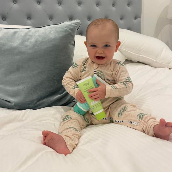 Joyful baby in bed holding Wotnot natural nappy rash cream